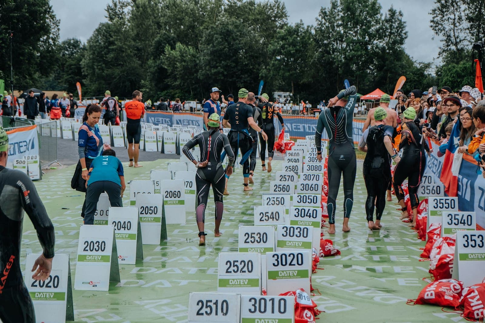 Triathletes in wetsuits gathered at the swim-start transition area on race morning, numbered transition bags lined up along the ground.